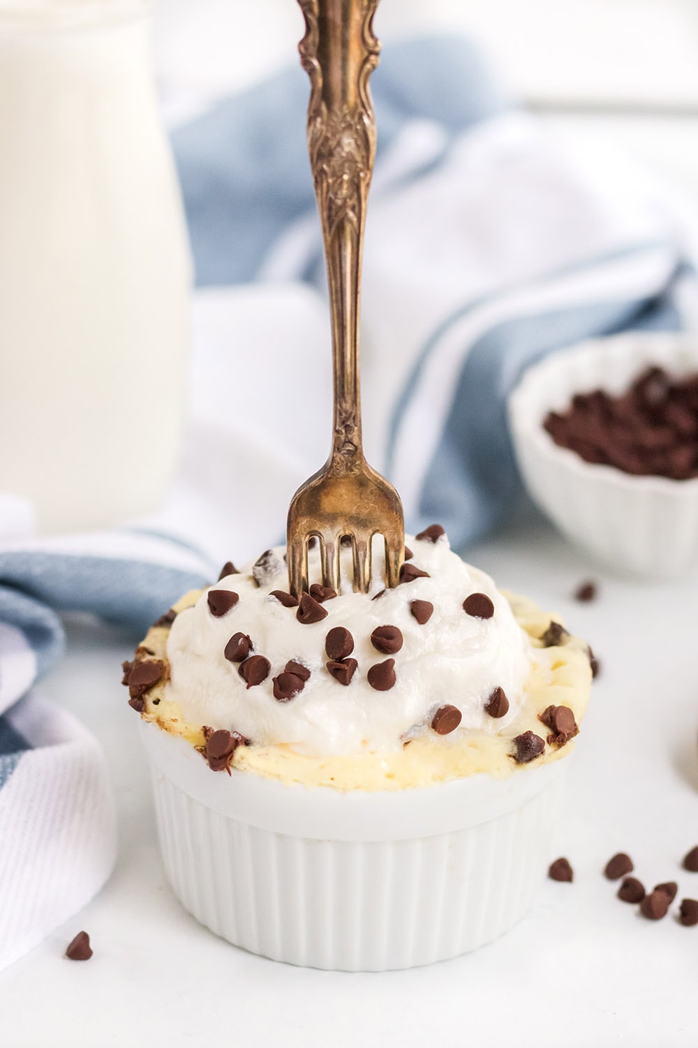 Fork in a chocolate chip mug cake surrounded by chocolate chips, blue napkin, and jar of milk.