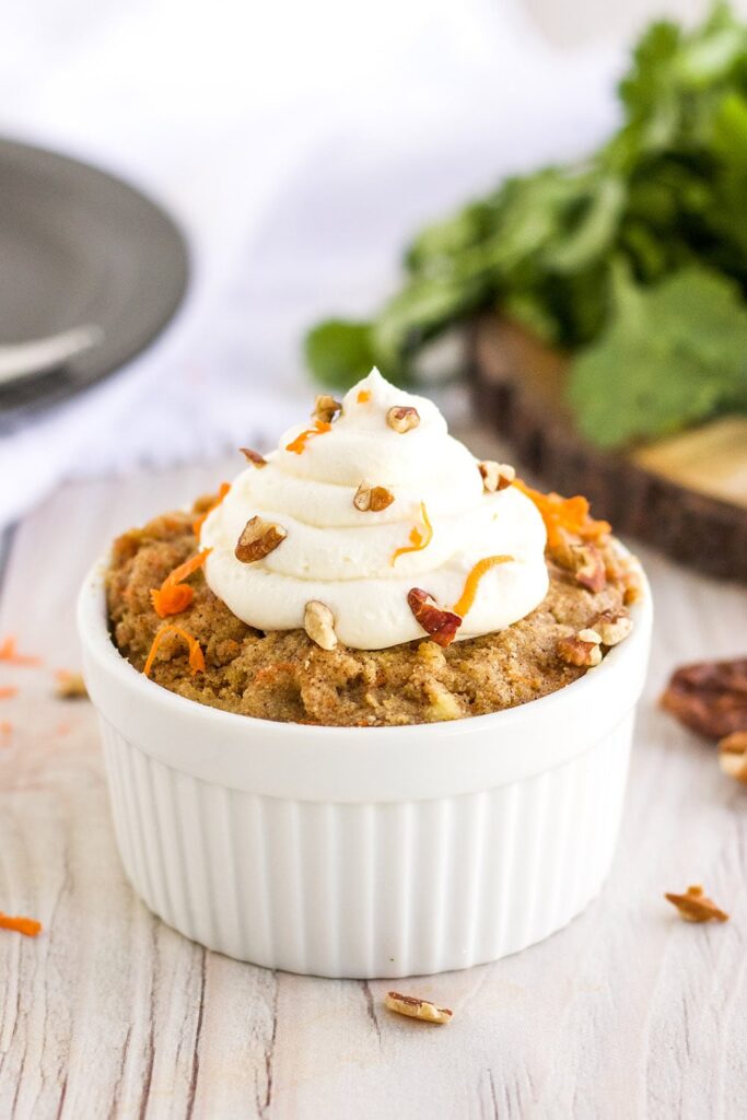Carrot cake topped with frosting, carrots, and pecans in a mug on a table