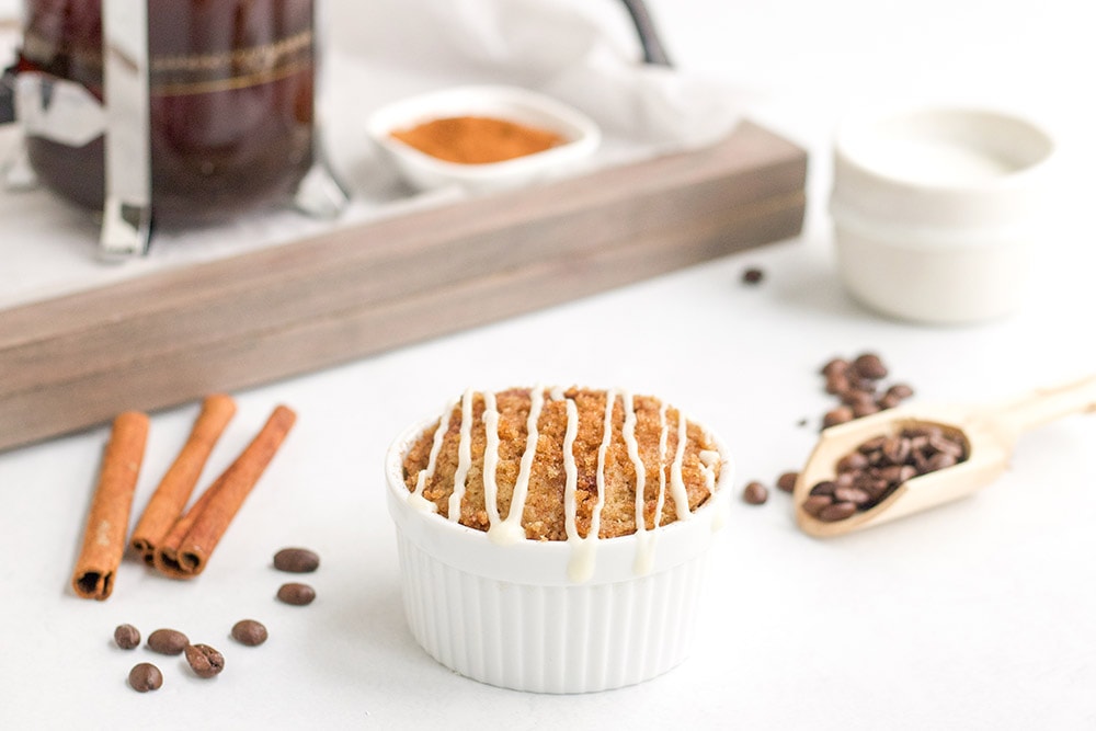 Coffee mug cake with glaze on top by a tray, coffee beans, and cinnamon sticks.