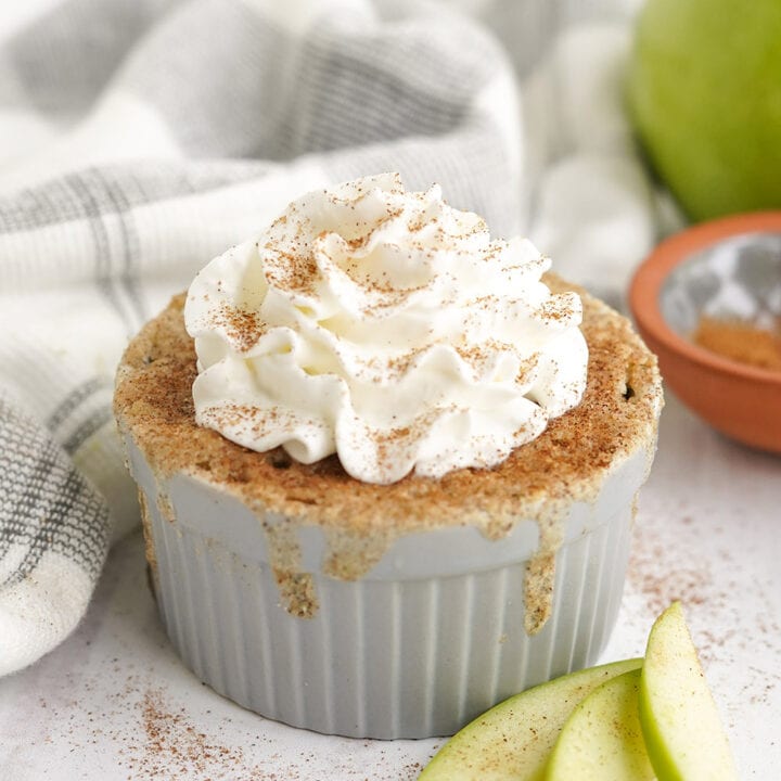 Cake topped with cinnamon and whipped cream in a gray dish with apple slices on the table.