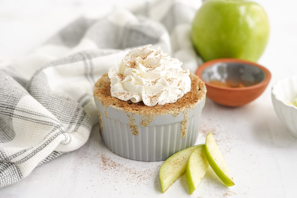 Apple cake in a ramekin topped with whipped cream and cinnamon. Apples and a napkin on the table.