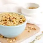 Banana cake in a mug topped with nuts and more chopped nuts on the table.