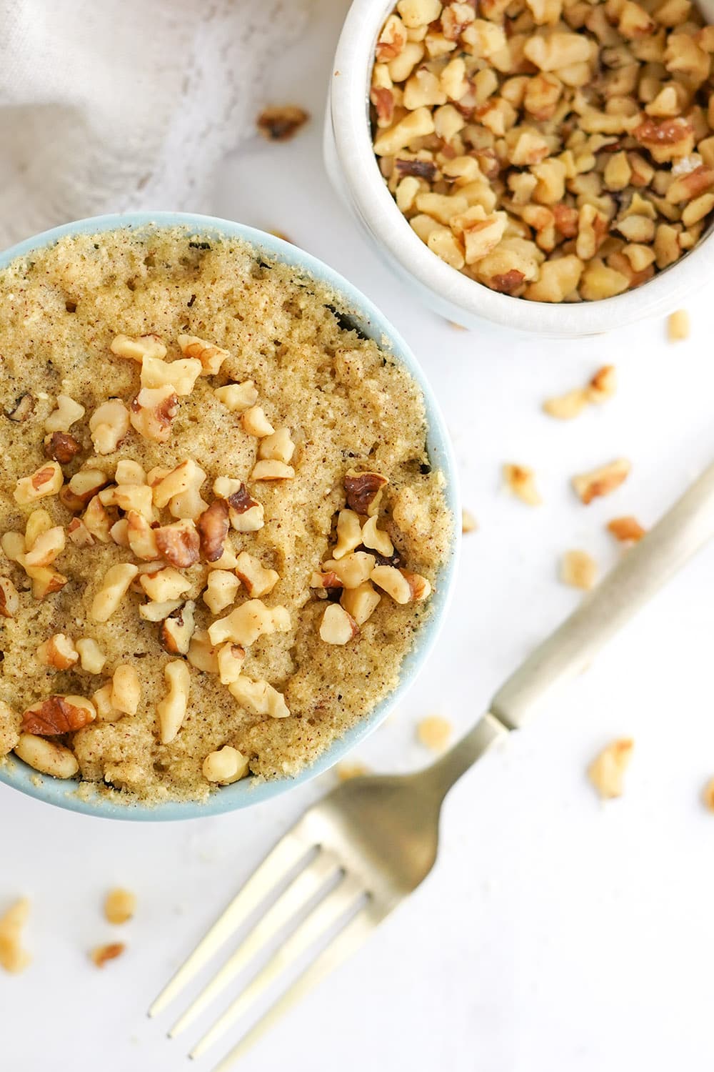 Overhead shot of banana cake in a mug with a bowl of chopped walnuts to the side.