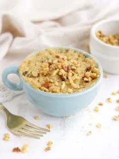 Banana cake in a mug with a fork and chopped walnuts on the table.