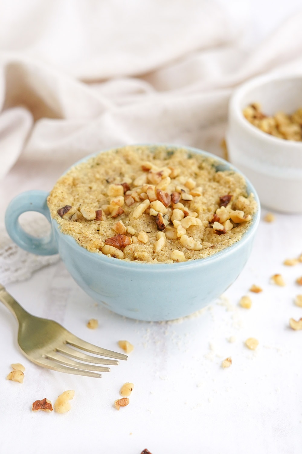 Banana cake in a mug with a fork and chopped walnuts on the table.