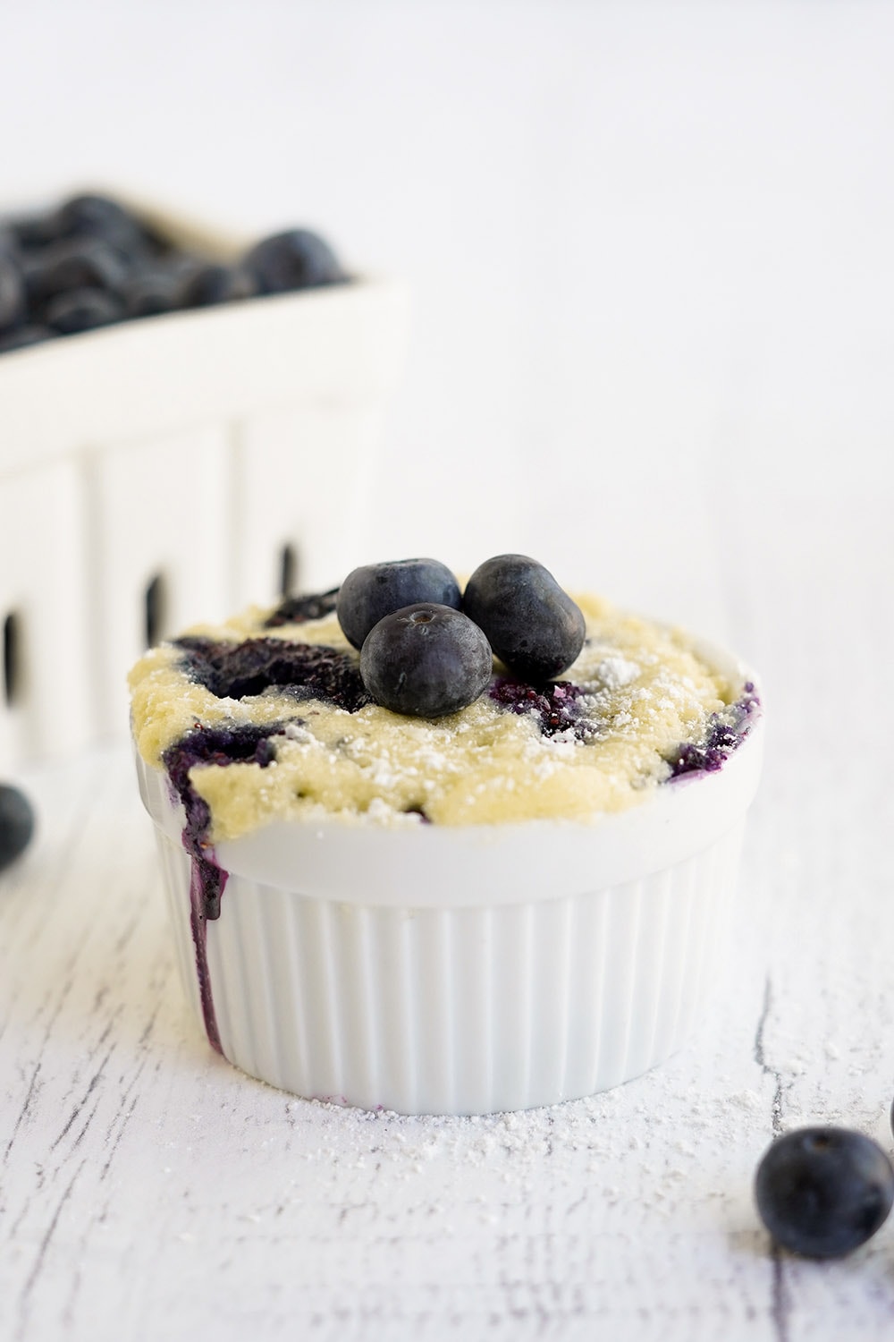 Blueberry cake in a mug with basket of berries on the table.