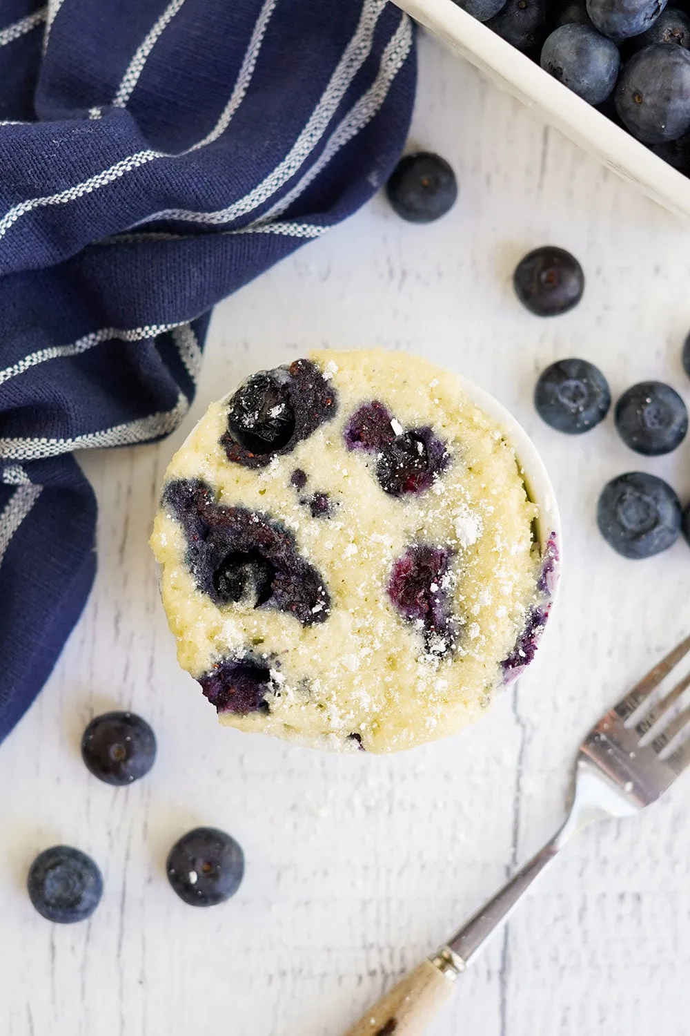Overhead shot of blueberry cake in a mug with a fork and more berries on the table.