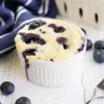 Blueberry cake in a mug with more berries on the table with a fork and navy and white striped napkin.