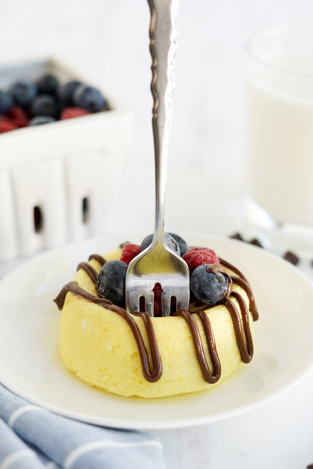 Fork standing in a cheesecake with chocolate and berries. Basket of blueberries in the background.