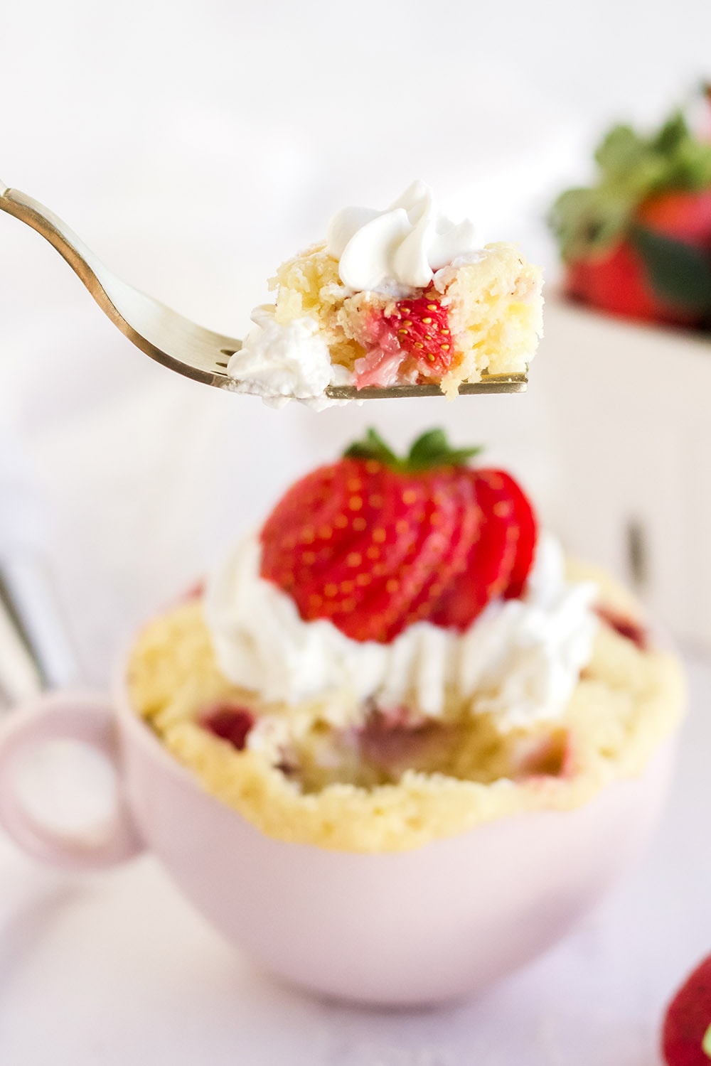 Fork with cake on it raised above strawberry cake in a mug.