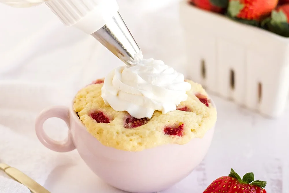Piping whipped cream on top of a strawberry cake in a pink mug.